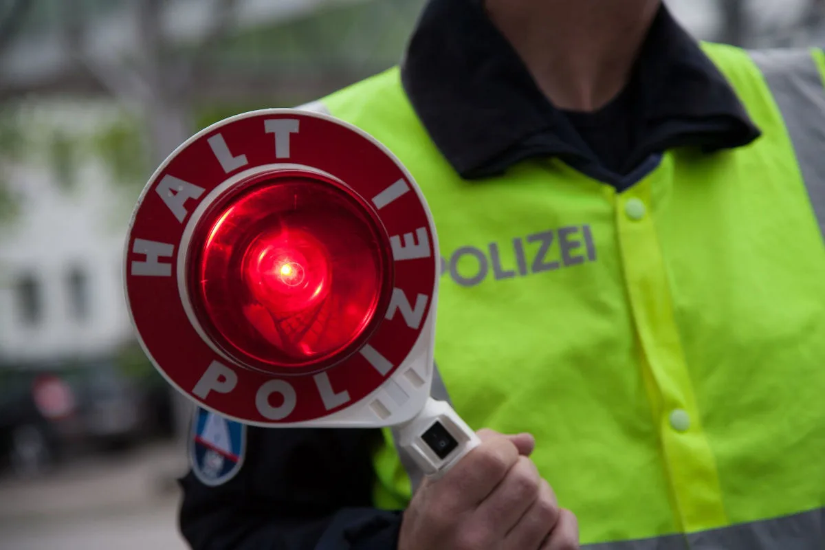 Ein Polizist hält eine rote Stoppscheibe mit der Aufschrift 'Halt Polizei'. Die Weste trägt den Schriftzug 'Polizei'.