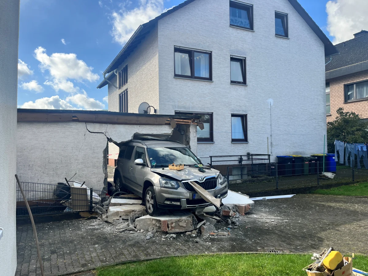 Ein silbernes Auto ist nach einem Unfall in eine Garagenwand gefahren, die dadurch teilweise eingestürzt ist. Im Hintergrund sind Wohnhäuser sichtbar.