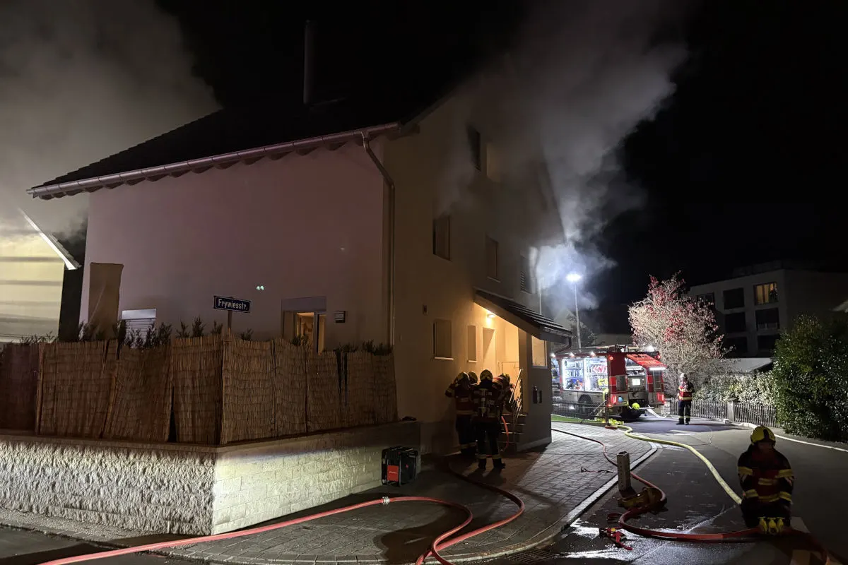Ein Wohnhaus zeigt starke Rauchentwicklung. Mehrere Feuerwehrleute sind im Einsatz. Ein großes Feuerwehrfahrzeug steht auf der Straße.