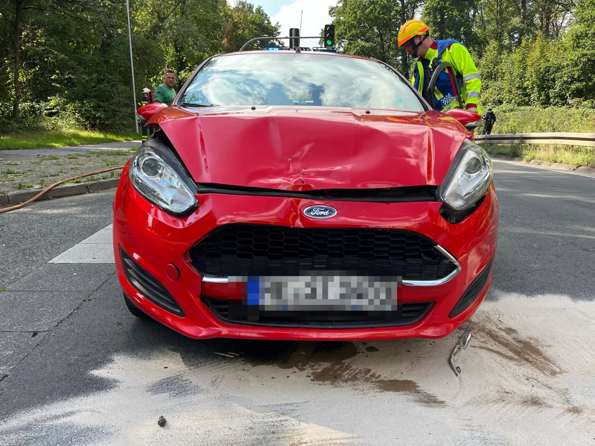 Ein roter Ford mit sichtbaren Frontschäden steht auf einer Straße. Im Hintergrund sind mehrere Personen und ein Feuerwehrmann zu sehen.