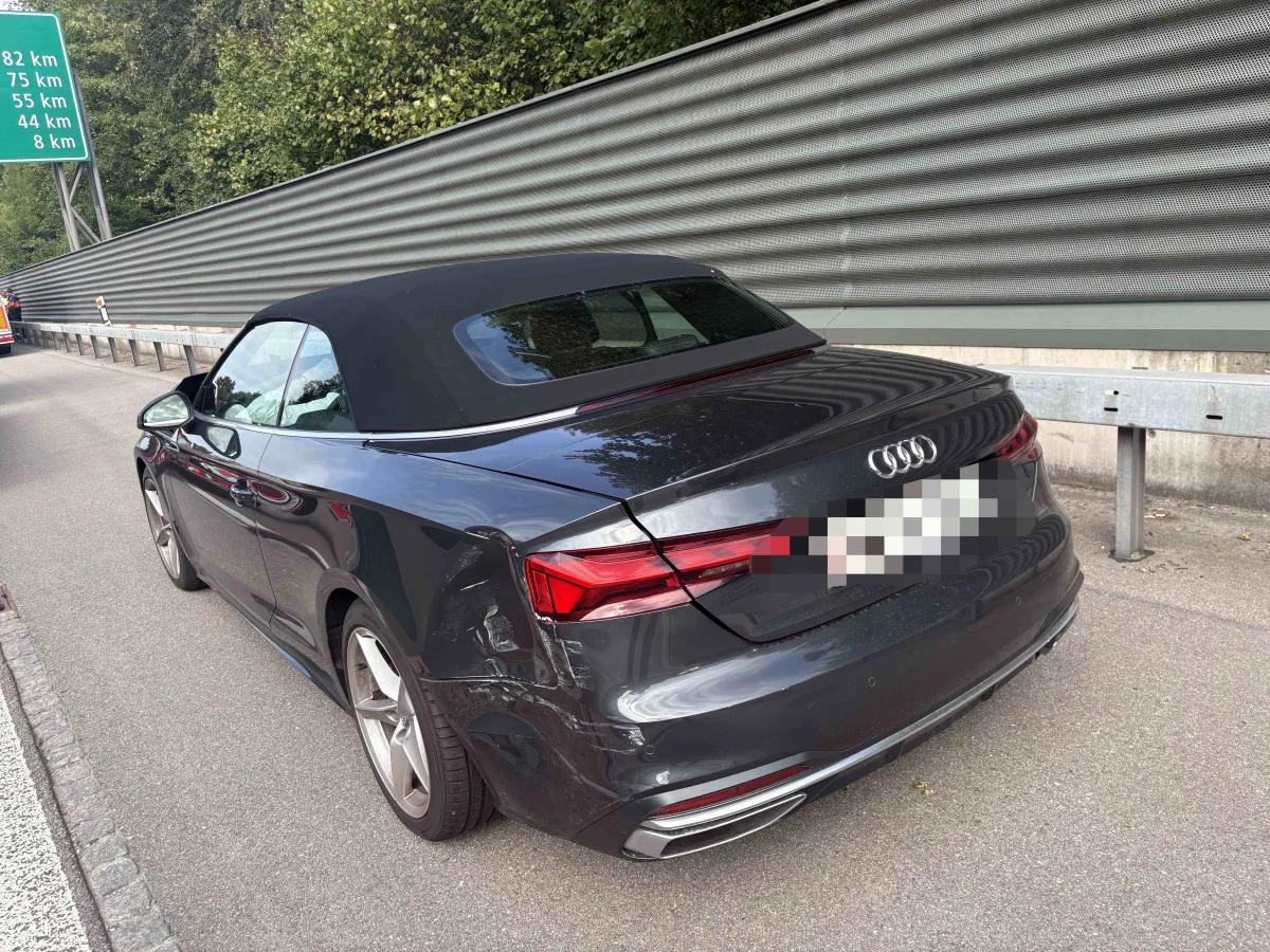 Ein dunkler Audi zeigt deutliche Kratzer am hinteren Kotflügel. Das Fahrzeug steht neben einer Leitplanke auf der Autobahn.