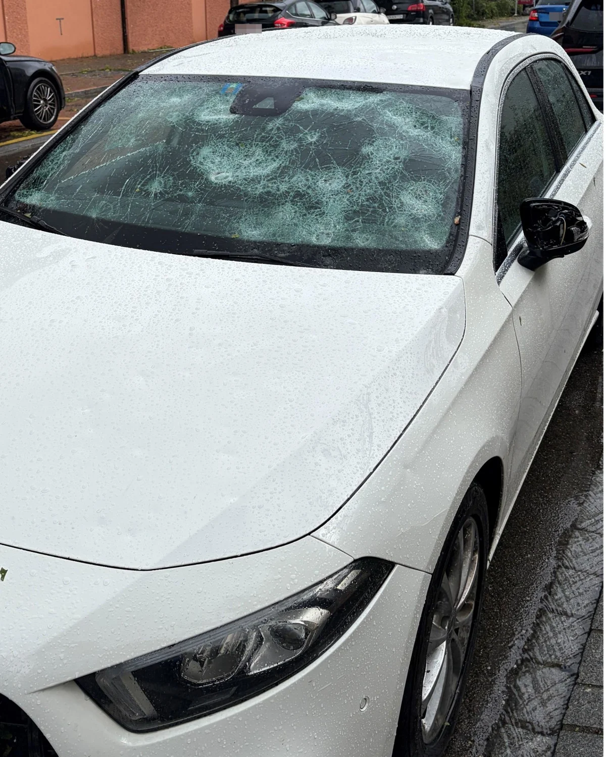 Ein weißes Auto steht am Straßenrand mit einer stark beschädigten Windschutzscheibe; es regnet.