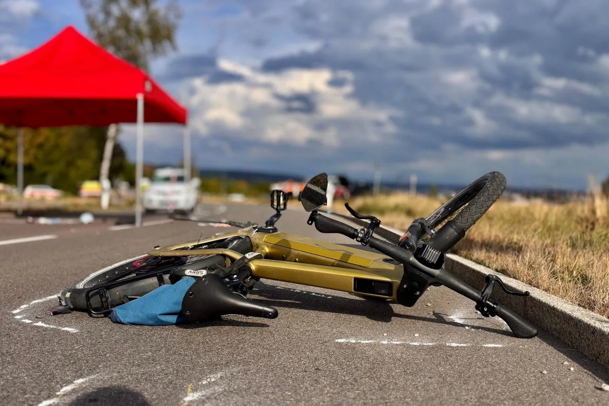 Ein goldfarbenes Fahrrad liegt auf einer asphaltierten Straße, im Hintergrund steht ein rotes Zelt. Der Himmel ist bewölkt.