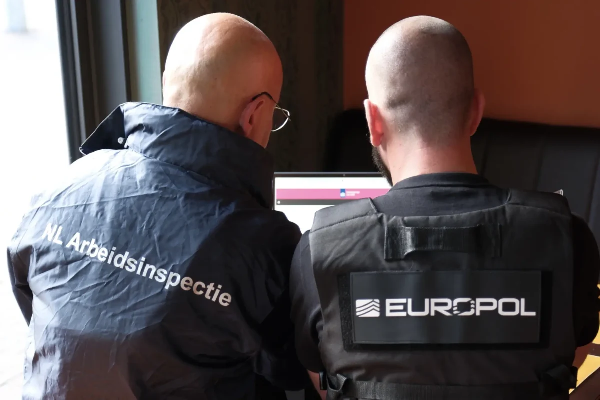 Zwei Männer in offizieller Bekleidung von 'NL Arbeidsinspectie' und 'Europol' sitzen vor einem Laptop, sichtbar von hinten.