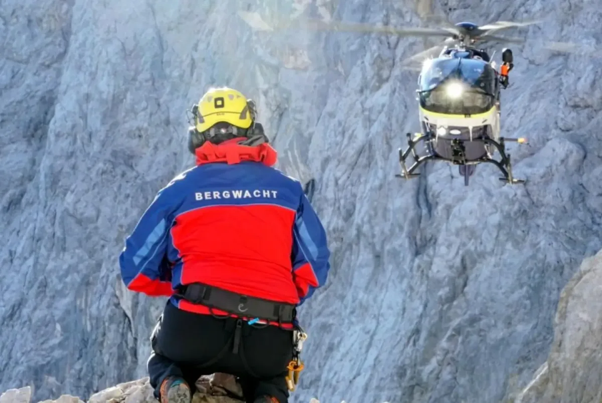 Ein Bergretter mit roter und blauer Kleidung und Helm sitzt auf Felsen, während ein Hubschrauber in der Luft schwebt.