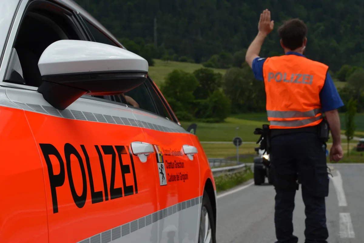 Ein Polizist in einer orangefarbenen Warnweste steht neben einem weißen Polizei-Fahrzeug mit orangefarbenen Markierungen. Im Hintergrund ist eine ländliche Straße zu sehen.