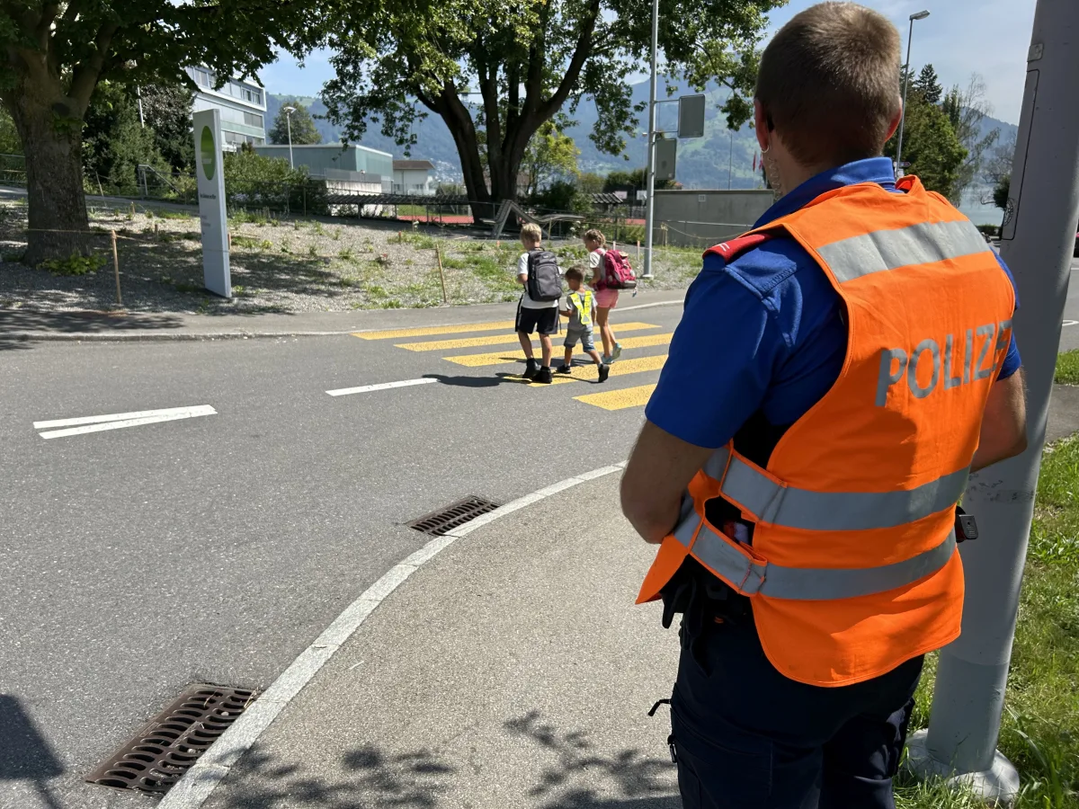 Ein Polizist in orangener Weste steht am Straßenrand und beobachtet drei Kinder, die einen Zebrastreifen überqueren.