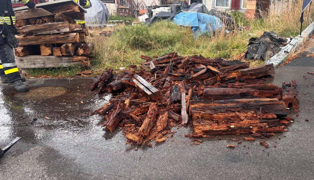 Verkohlte Holzstücke liegen auf nassem Asphalt. Im Hintergrund sind Feuerwehrleute zu sehen.