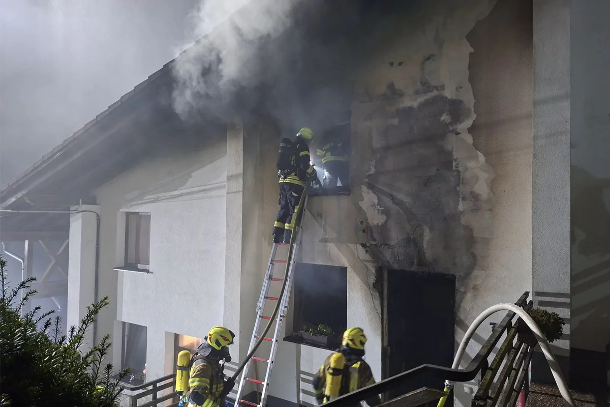 Mehrere Feuerwehrleute mit Atemschutzmasken bekämpfen einen Brand an einem Fenster eines stark verrußten Hauses. Dicker Rauch dringt aus dem Gebäude, während sie auf einer Leiter stehen.
