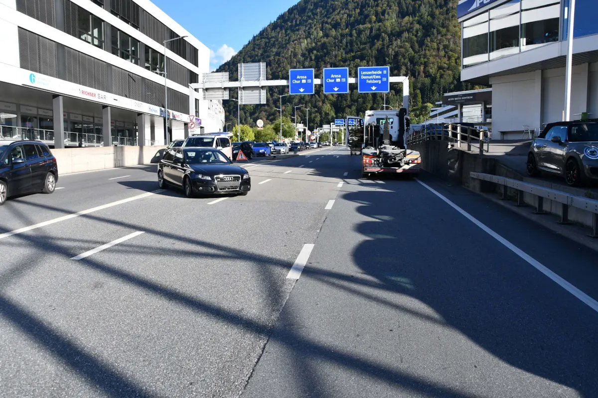 Eine befahrene Straße in Chur zeigt mehrere Fahrzeuge. Verkehrszeichen weisen auf Ziele wie Arosa und Chur hin.