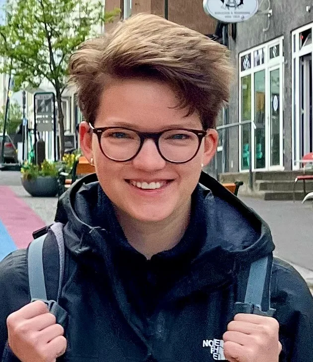 Eine lächelnde Person mit Brille und kurzem Haar trägt eine Jacke und einen Rucksack und steht auf einer farbigen Straße.