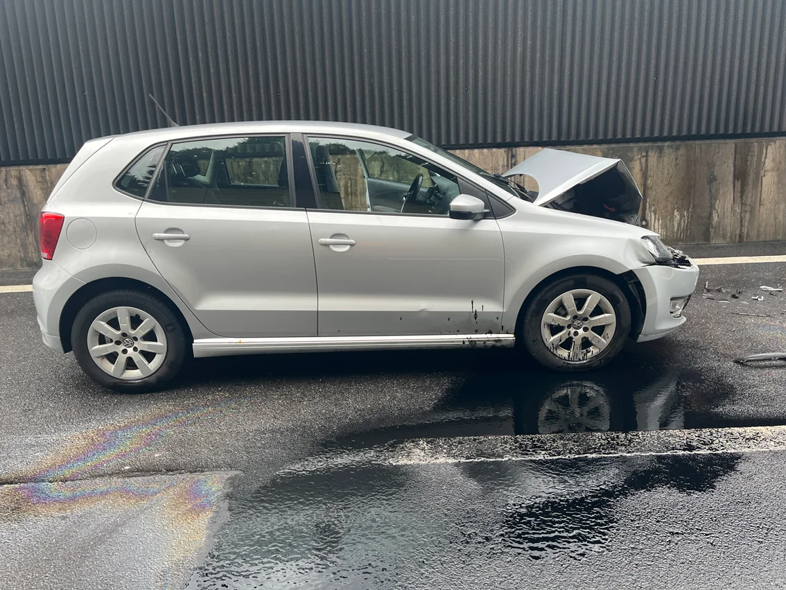 Ein silbernes Auto steht mit geöffnetem Motorhaube und sichtbaren Schäden auf einer nassen Straße. Regenbogenfarbige Ölflecken sind auf dem Boden zu sehen.