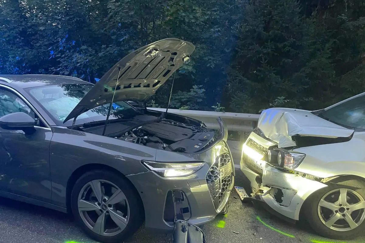 Ein grauer Audi und ein weißes Auto stehen nach einem Unfall aneinandergestoßen auf einer Straße. Der Audi hat die Motorhaube geöffnet.