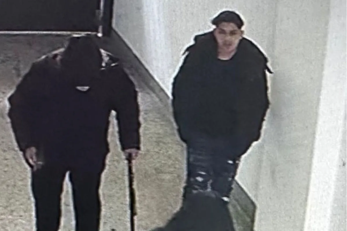 Zwei Männer gehen durch einen hellen Flur. Der linke Mann trägt einen Stock und hat eine Kapuze auf.