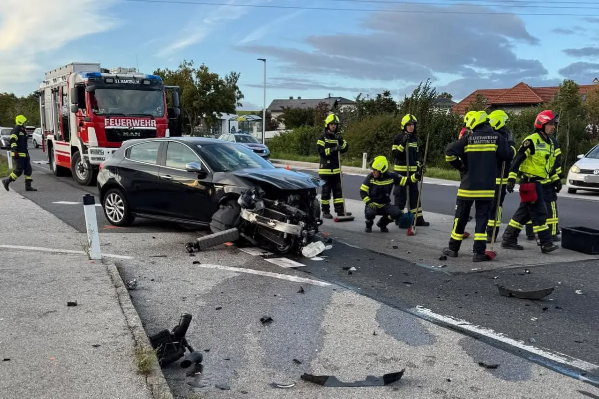 Ein schwarzes Auto ist nach einem Unfall stark beschädigt, umgeben von Feuerwehrleuten in Schutzkleidung auf der Straße.