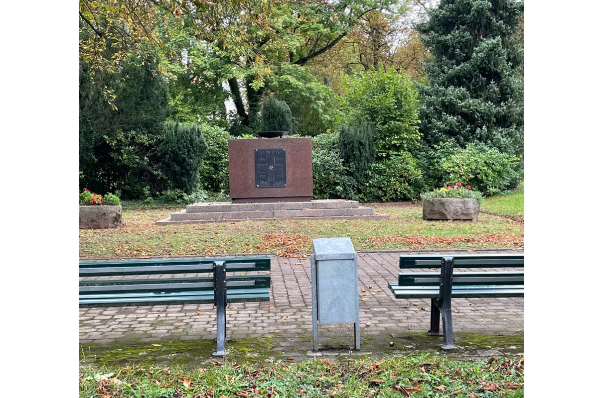 Ein Denkmal aus Stein steht in einem grünen Park. Im Vordergrund sind zwei Bänke und ein Mülleimer zu sehen, umgeben von Bäumen und Sträuchern.