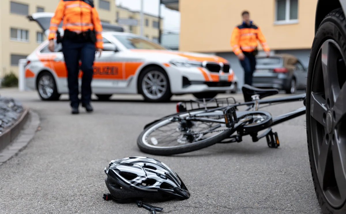 Ein umgestürztes Fahrrad und ein Fahrradhelm liegen auf einer Straße vor einem weißen Polizeiauto mit orangenen Markierungen. Zwei Polizisten in orangefarbenen Jacken stehen im Hintergrund.