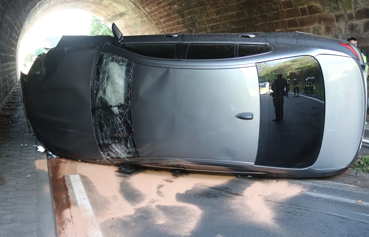 Ein silberfarbenes Auto liegt seitlich gekippt in einem Tunnel. Die Fahrbahn ist teilweise blockiert und es sind einige Personen am Rand sichtbar.