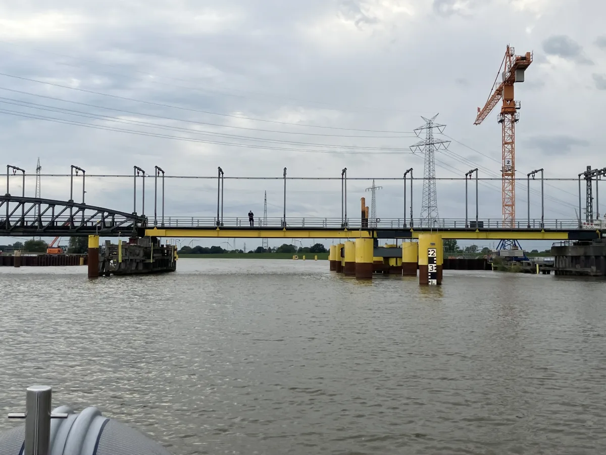 Eine Brücke überquert einen Fluss, darauf steht eine Person. Im Hintergrund ist ein großer Baukran zu sehen.
