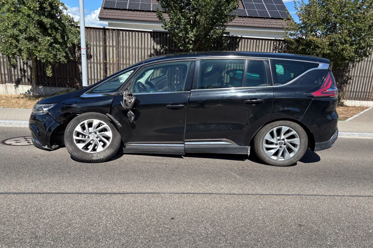 Ein schwarzes Auto mit sichtbarem Seitenschaden steht auf einer asphaltierten Straße bei klarem Wetter. Im Hintergrund sind Bäume und ein Zaun sichtbar.