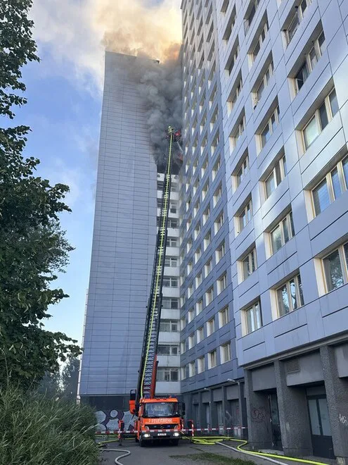 Ein Feuerwehrfahrzeug mit ausgefahrener Drehleiter bekämpft einen Brand in einem Hochhaus. Rauch steigt aus den oberen Etagen auf.