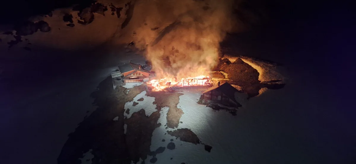 Ein Gebäude in alpiner Umgebung steht nachts in Flammen. Dichte Rauchschwaden steigen auf, umgeben von schneebedeckten Flächen.