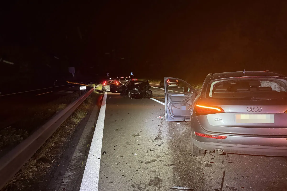 Mehrere Fahrzeuge stehen beschädigt auf einer dunklen Autobahn. Trümmerteile liegen verstreut, und ein Fahrzeug hat die Tür geöffnet.