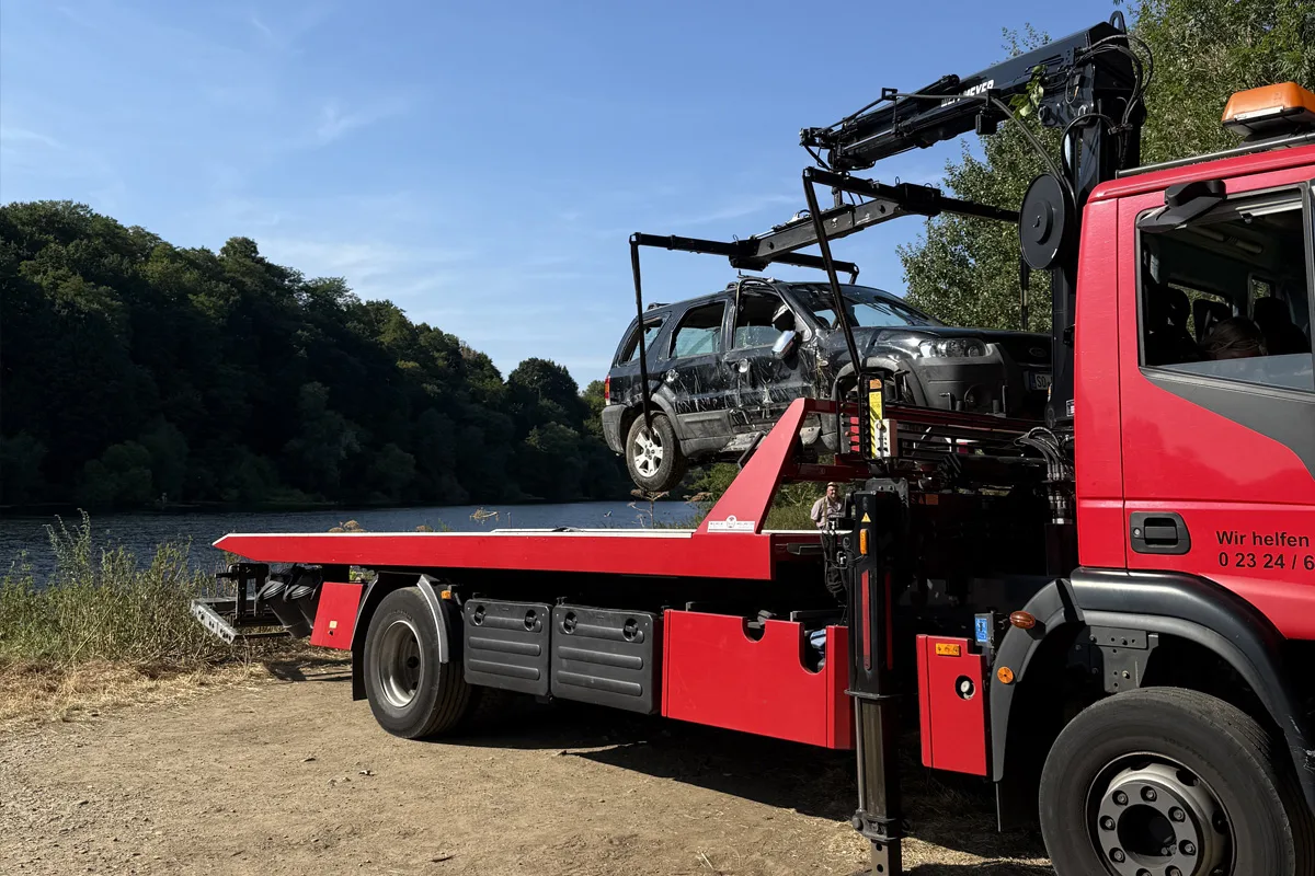 Ein roter Abschleppwagen hebt ein beschädigtes Auto an einem Flussufer. Das Fahrzeug zeigt deutliche Unfallspuren.