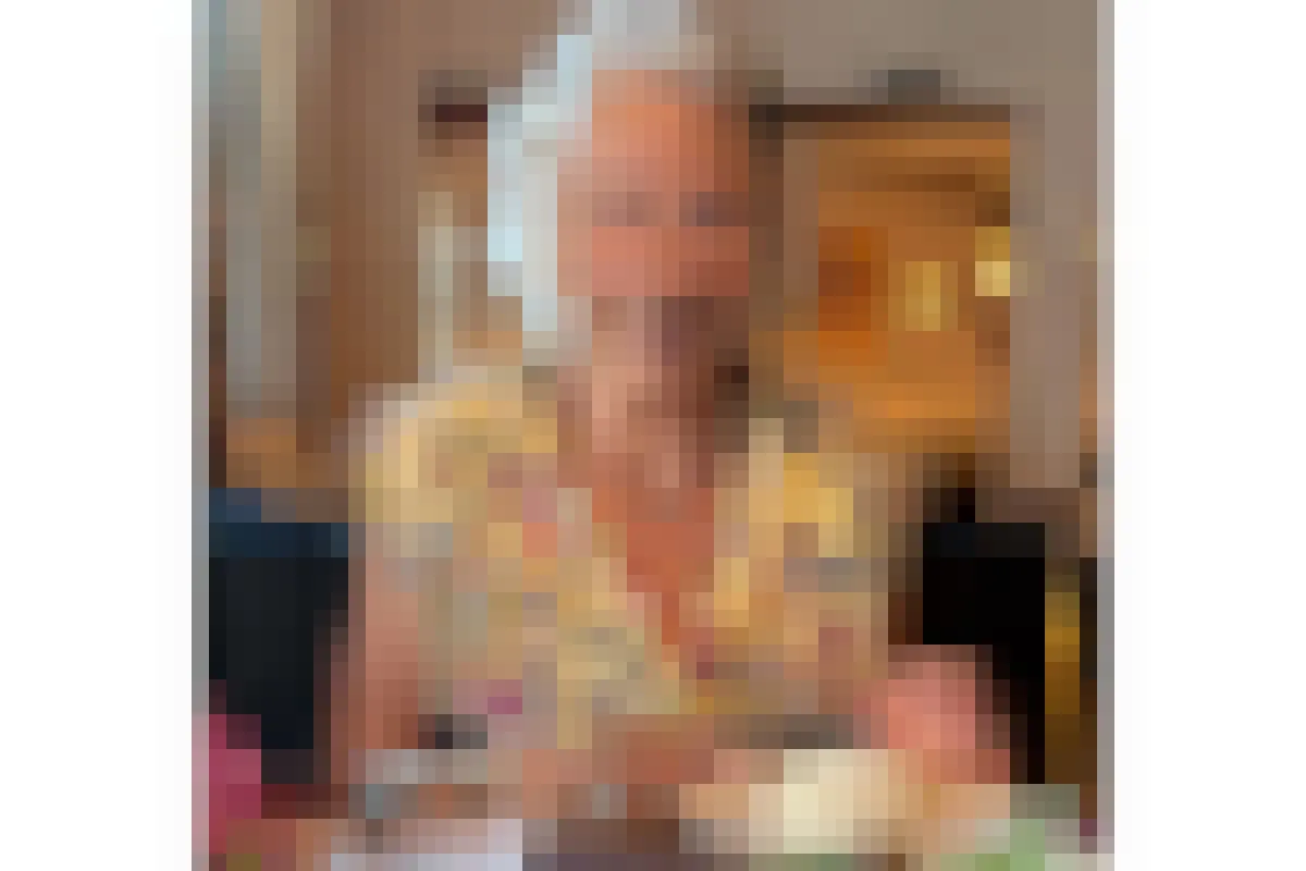 Eine ältere Dame mit grauen Haaren sitzt an einem Tisch in einem Innenraum. Vor ihr steht ein Dessert auf dem Tisch.