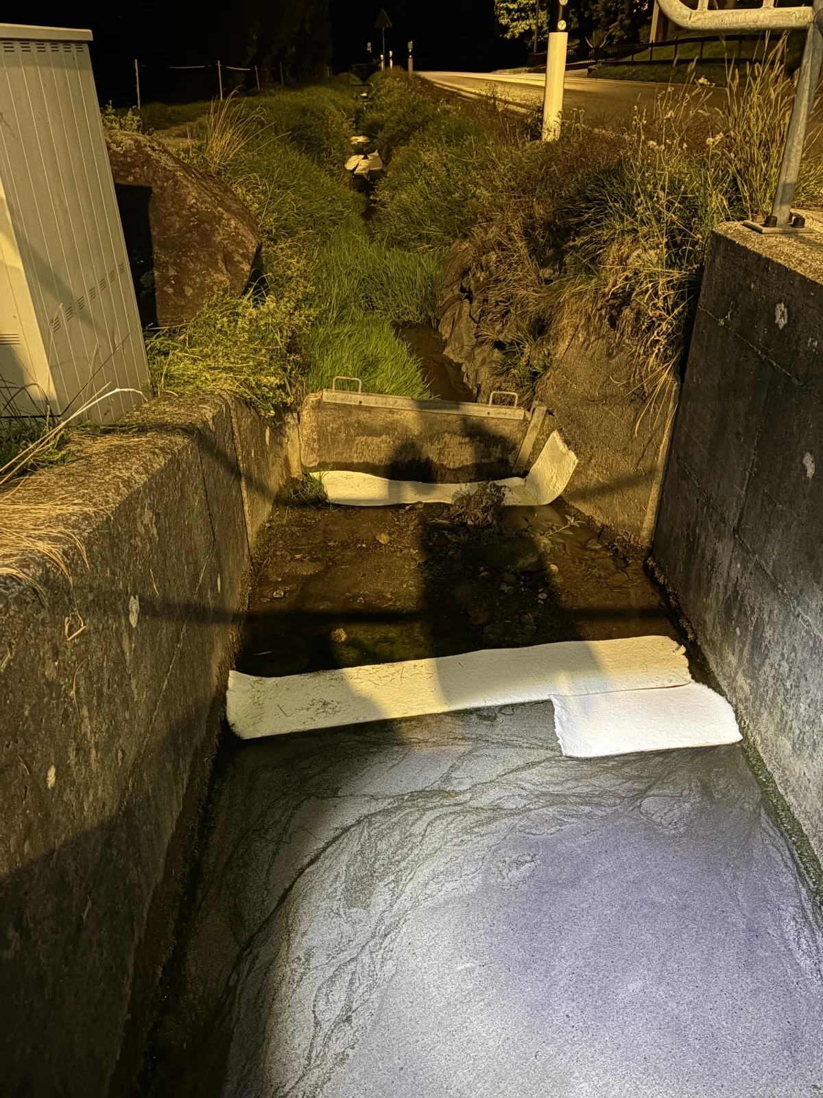 Ein Entwässerungsgraben bei Nacht, teilweise blockiert durch weißes Material, im Scheinwerferlicht beleuchtet.