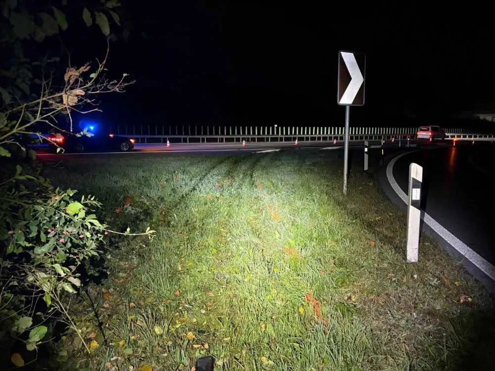 Nachtaufnahme einer Landstraße mit einem Kurvenschild, Verkehrskegeln und Fahrzeugen im Hintergrund, die die Straße beleuchten.