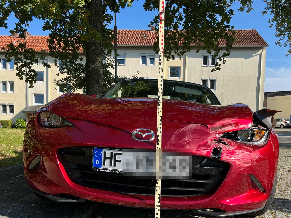 Ein roter Mazda mit erheblichen Schäden an der Front steht auf einem Parkplatz. Im Hintergrund sind Wohngebäude sichtbar.
