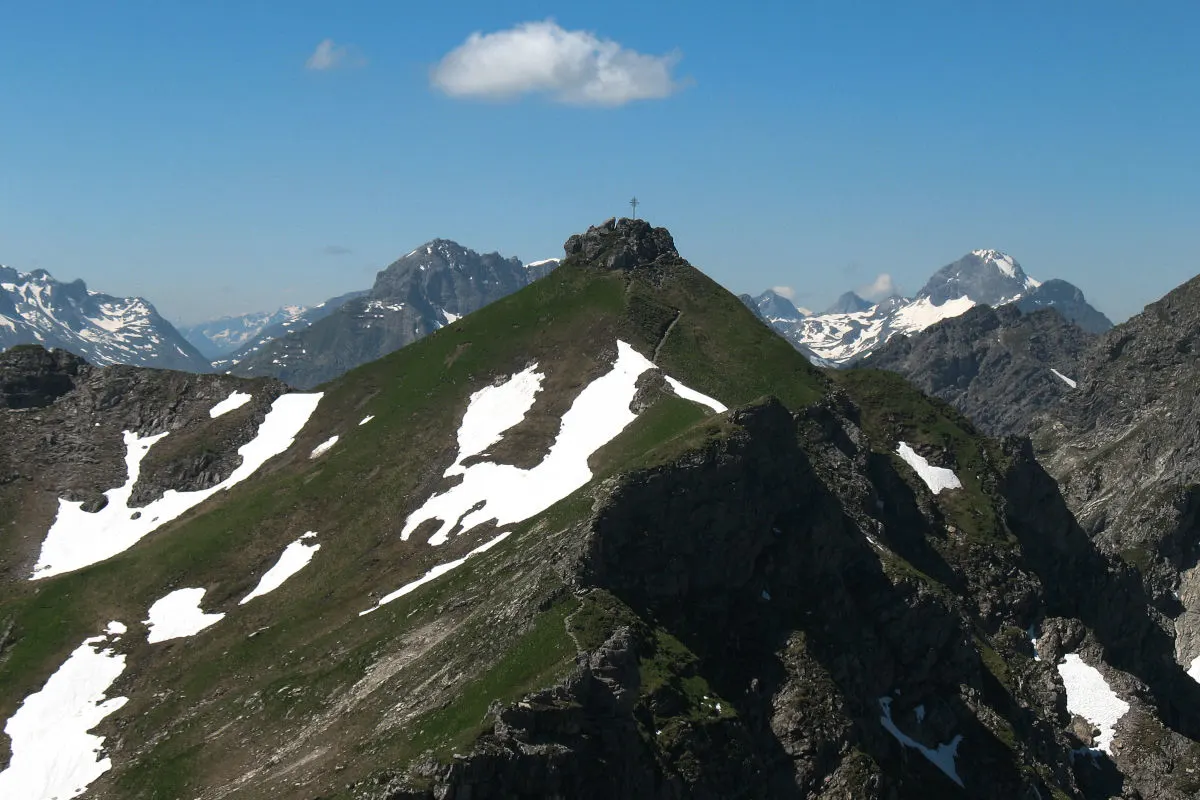 Ein Gipfel in den Alpen ist teilweise mit Schnee bedeckt, während ein Kreuz auf dem höchsten Punkt steht. Der Himmel ist klar mit wenigen Wolken.