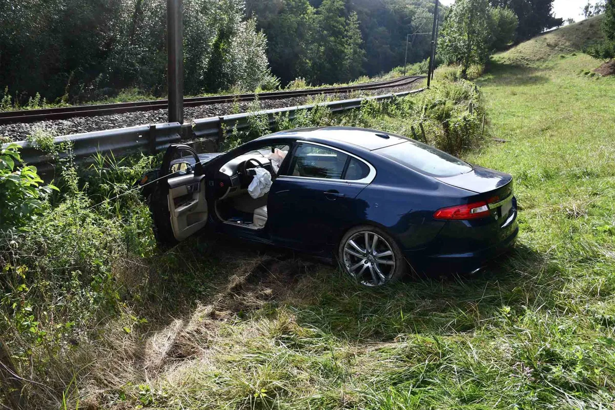 Ein blaues Auto steht mit geöffneter Tür und ausgelöstem Airbag neben Bahngleisen. Die Umgebung ist grasbewachsen und ländlich.