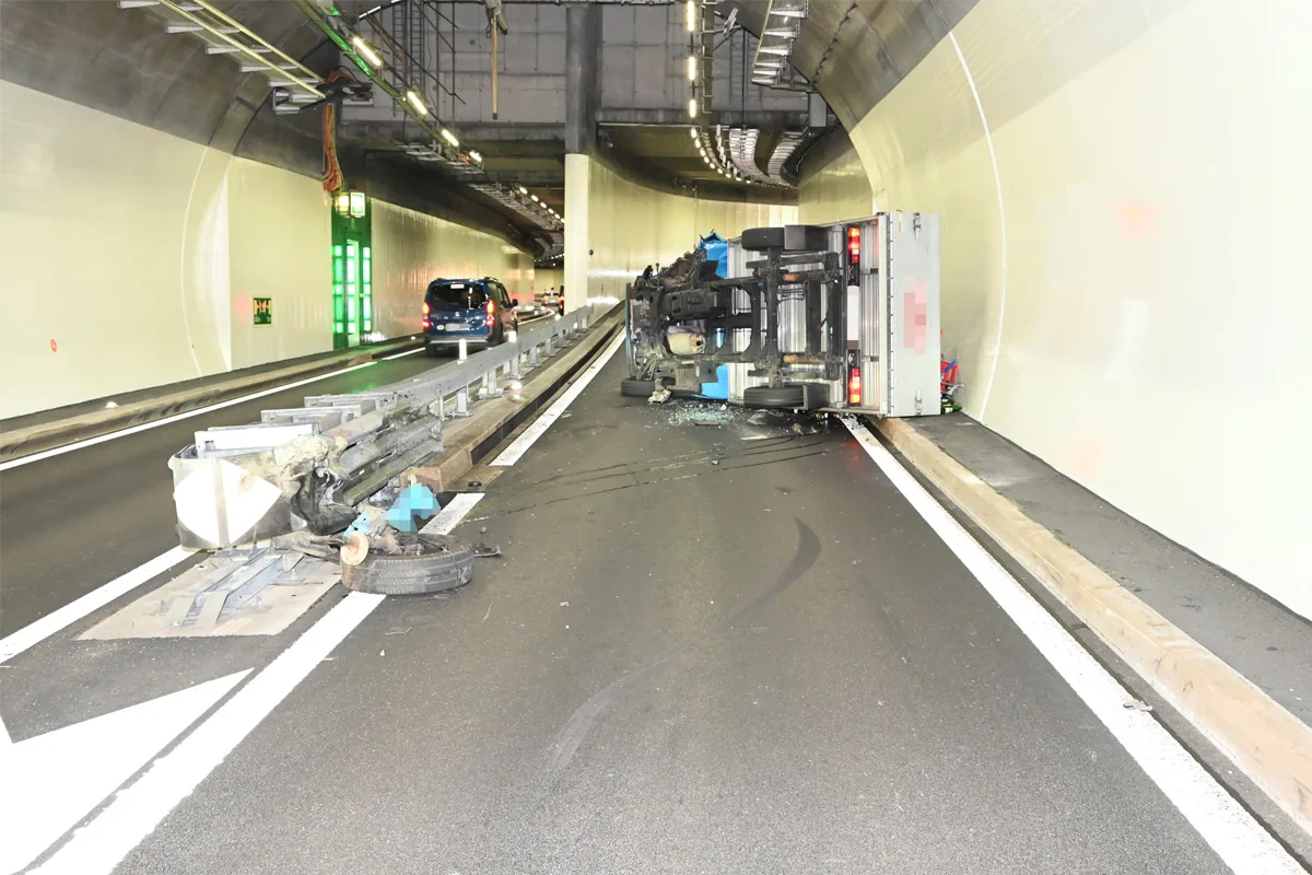 Ein Lastwagen liegt seitlich in einem Tunnel und blockiert eine Fahrbahnhälfte. Autos scheinen auf der freien Spur vorbeizufahren. Trümmer liegen verstreut auf dem Boden.