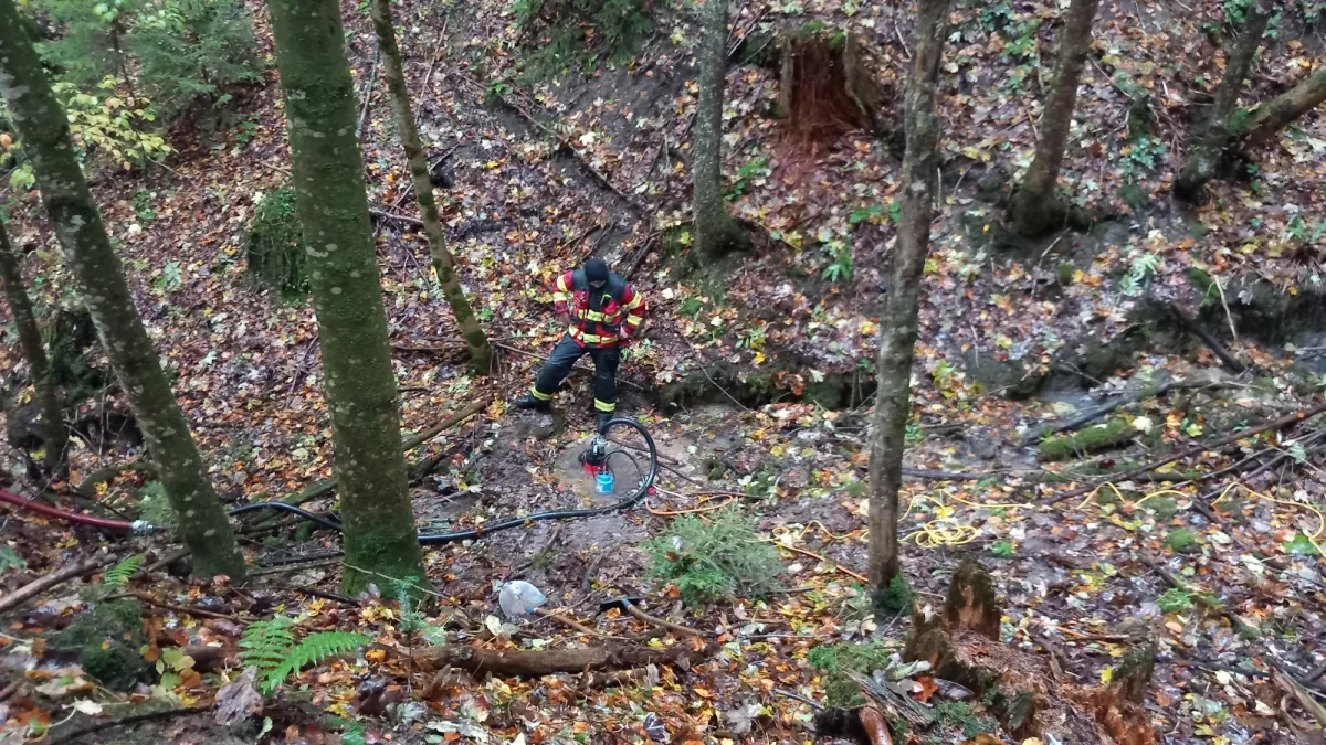 Ein Feuerwehrmann in Uniform steht im Wald und ist mit einer Pumpe beschäftigt, umgeben von Laub auf dem Boden.