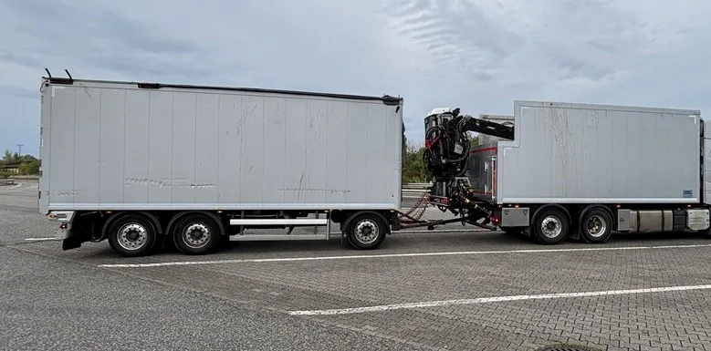 Zwei große weiße Lastwagen sind auf einem leeren Parkplatz miteinander verbunden. Der Himmel wirkt bewölkt.