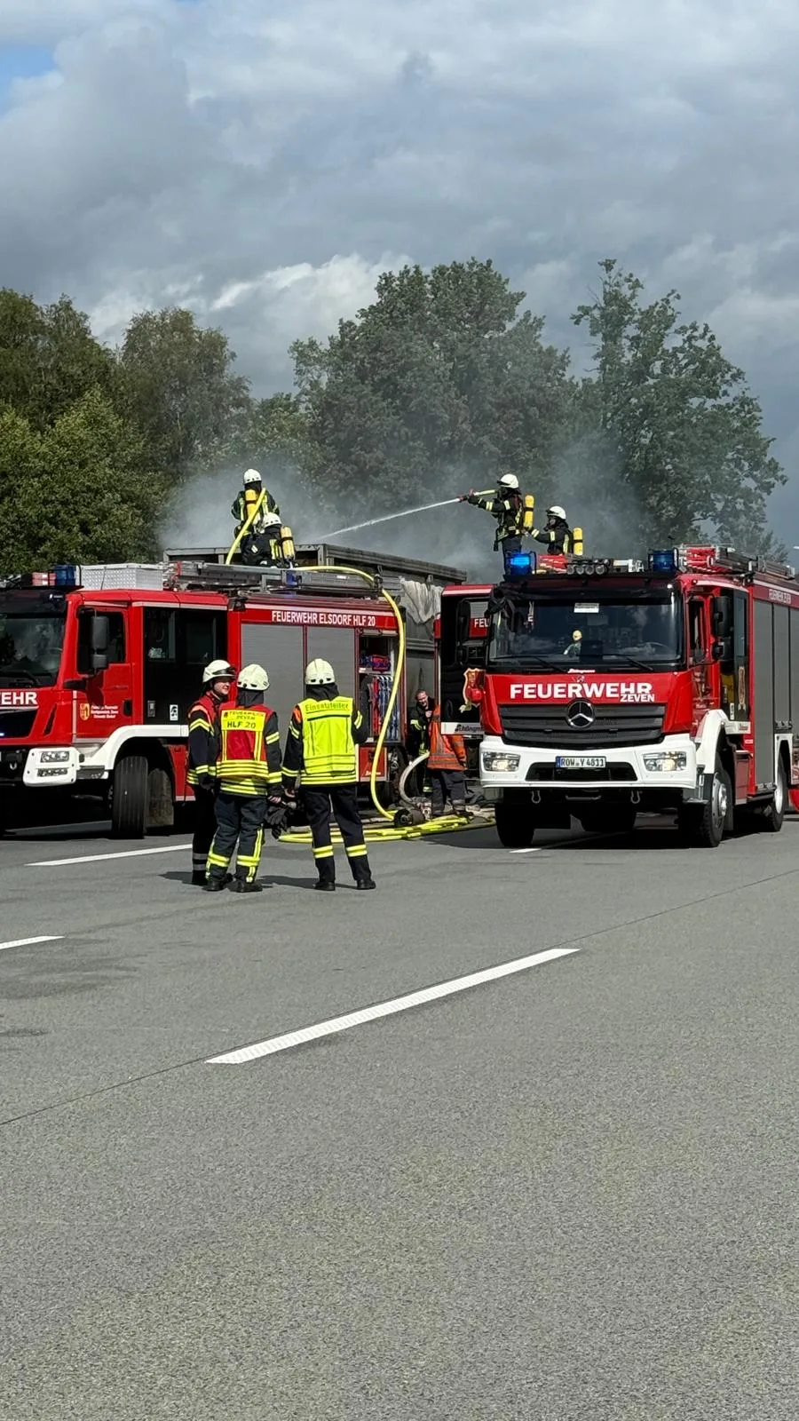 Mehrere Feuerwehrleute sind im Einsatz, um ein brennendes Fahrzeug auf einer Straße zu löschen. Rauch steigt aus dem Fahrzeug auf.