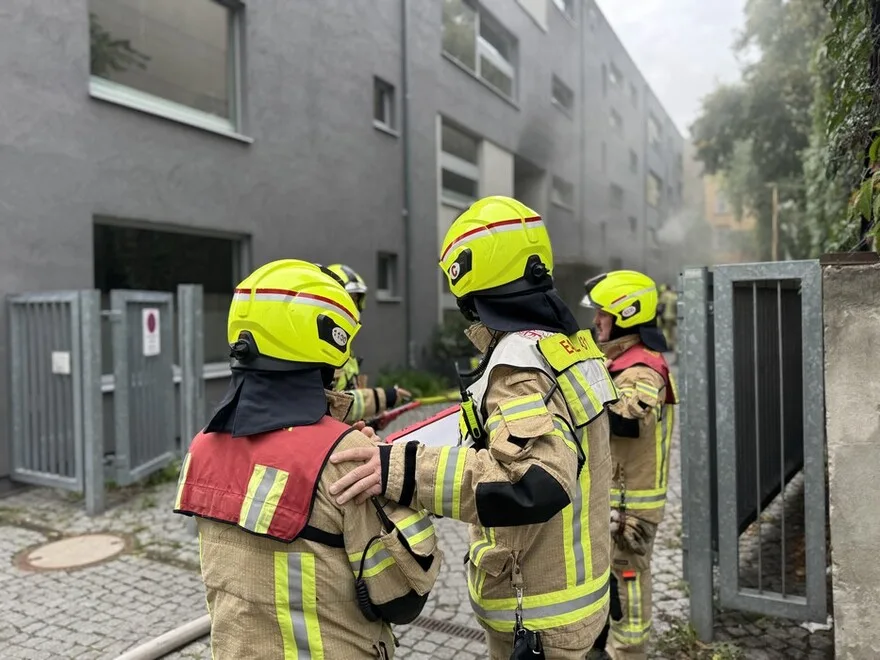 Mehrere Feuerwehrleute mit gelben Helmen stehen vor einem grauen Wohngebäude. Rauch ist im Hintergrund sichtbar.