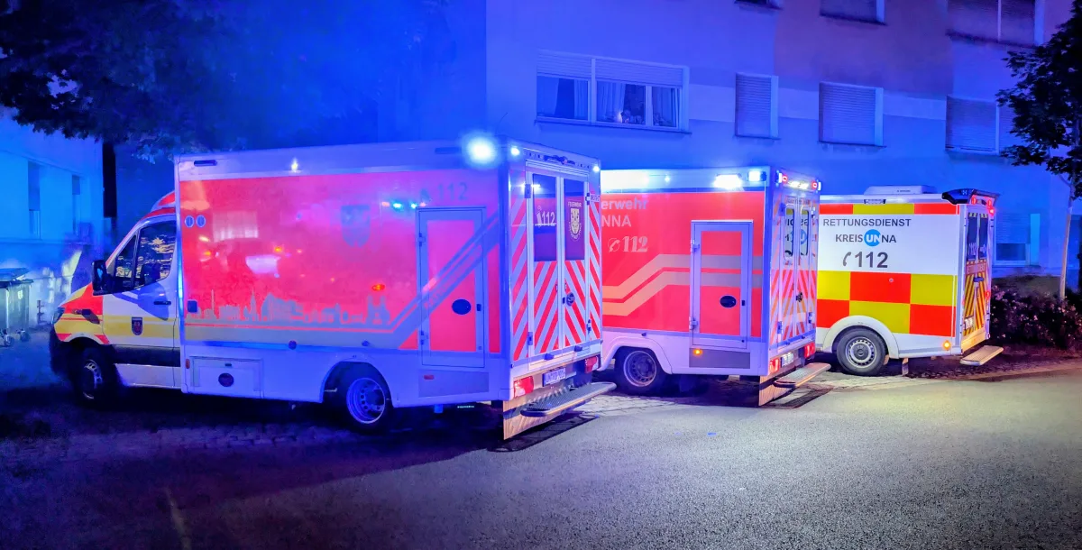 Die Szene zeigt drei rot-gelbe Rettungswagen mit aktiviertem Blaulicht geparkt vor einem Wohnhaus bei Nacht. Die Fahrzeuge tragen die Aufschrift 'Rettungsdienst Kreis Unna' und sind mit Notrufnummern gekennzeichnet.