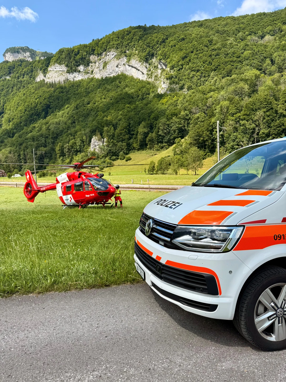 Ein roter Rettungshubschrauber steht auf einer grünen Wiese, flankiert von einem Polizeiauto. Im Hintergrund sind bewaldete Berge zu sehen.
