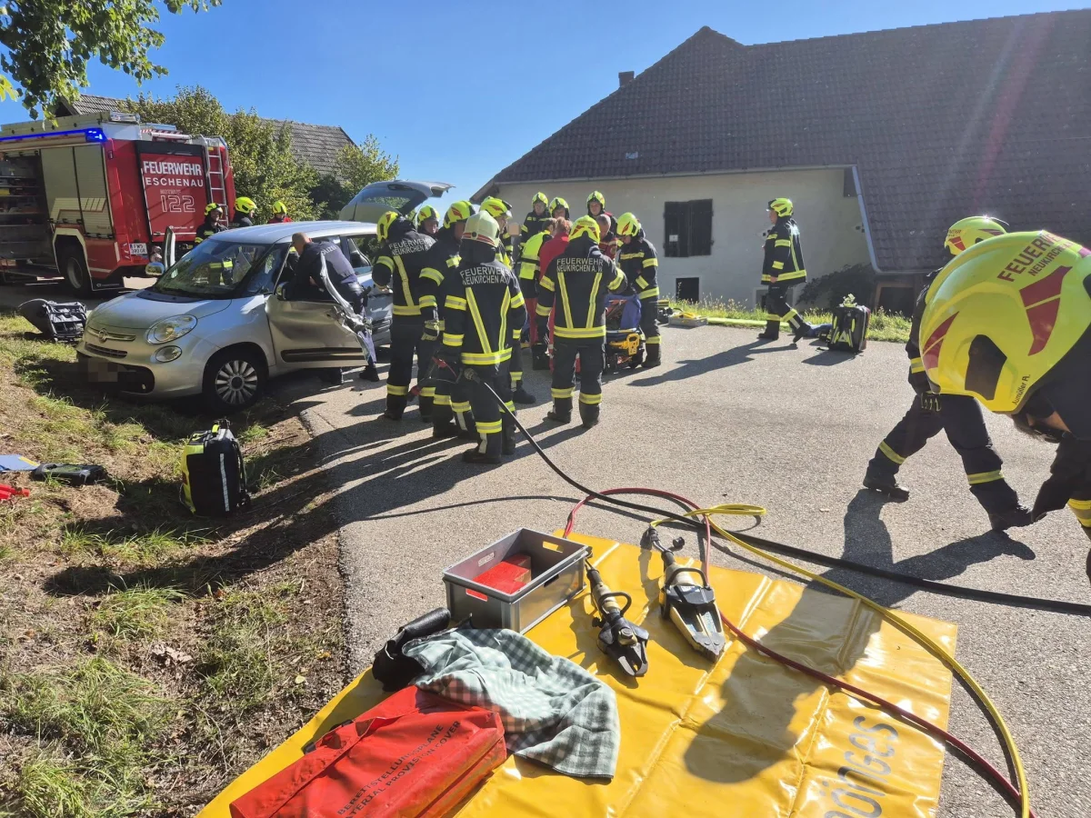 Mehrere Feuerwehrleute in Einsatzkleidung umgeben ein silbernes Auto auf einer Landstraße bei einem Haus. Ein Feuerwehrfahrzeug steht in der Nähe.