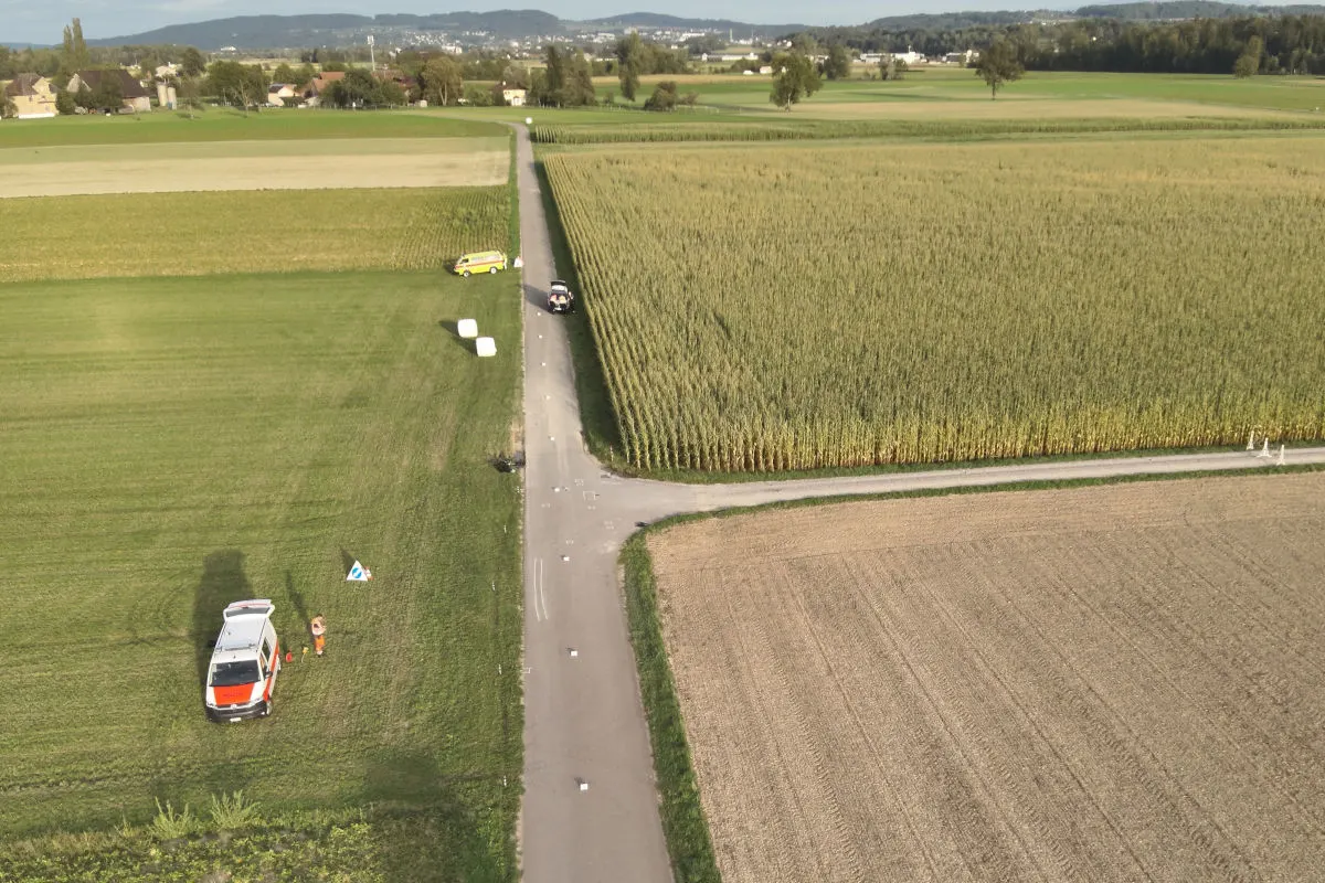 Einige Fahrzeuge stehen entlang einer schmalen Landstraße, die durch weitläufige Felder führt. Ein Rettungsfahrzeug ist auf einer Wiese geparkt.