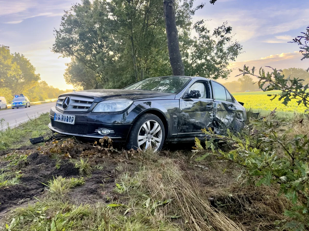 Ein beschädigter schwarzer Mercedes ist von der Straße abgekommen und steht neben einem Baum. Im Hintergrund sind Polizei- und Rettungsfahrzeuge zu sehen.