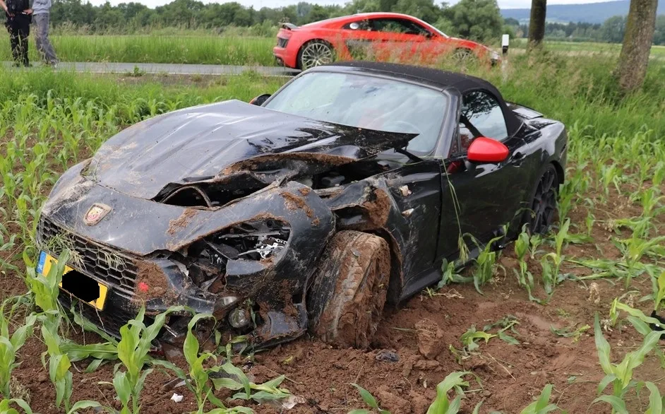 Ein schwarzer Sportwagen mit schweren Frontschäden steckt im aufgeweichten Boden eines Feldes fest. Im Hintergrund steht ein weiteres rotes Auto am Straßenrand.