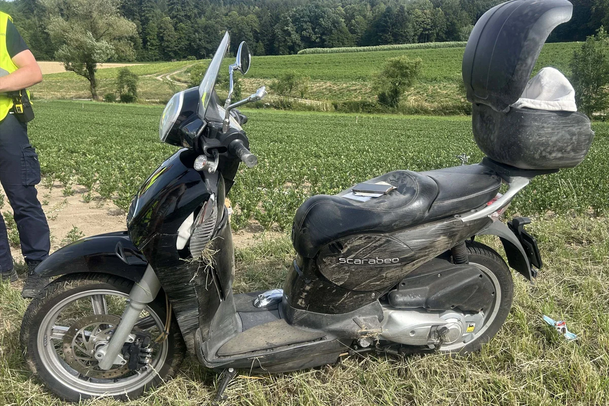 Ein schwarzer Motorroller mit Kratzern und Schmutz steht auf einem Feldweg. Daneben steht ein Polizist mit einer gelben Weste.