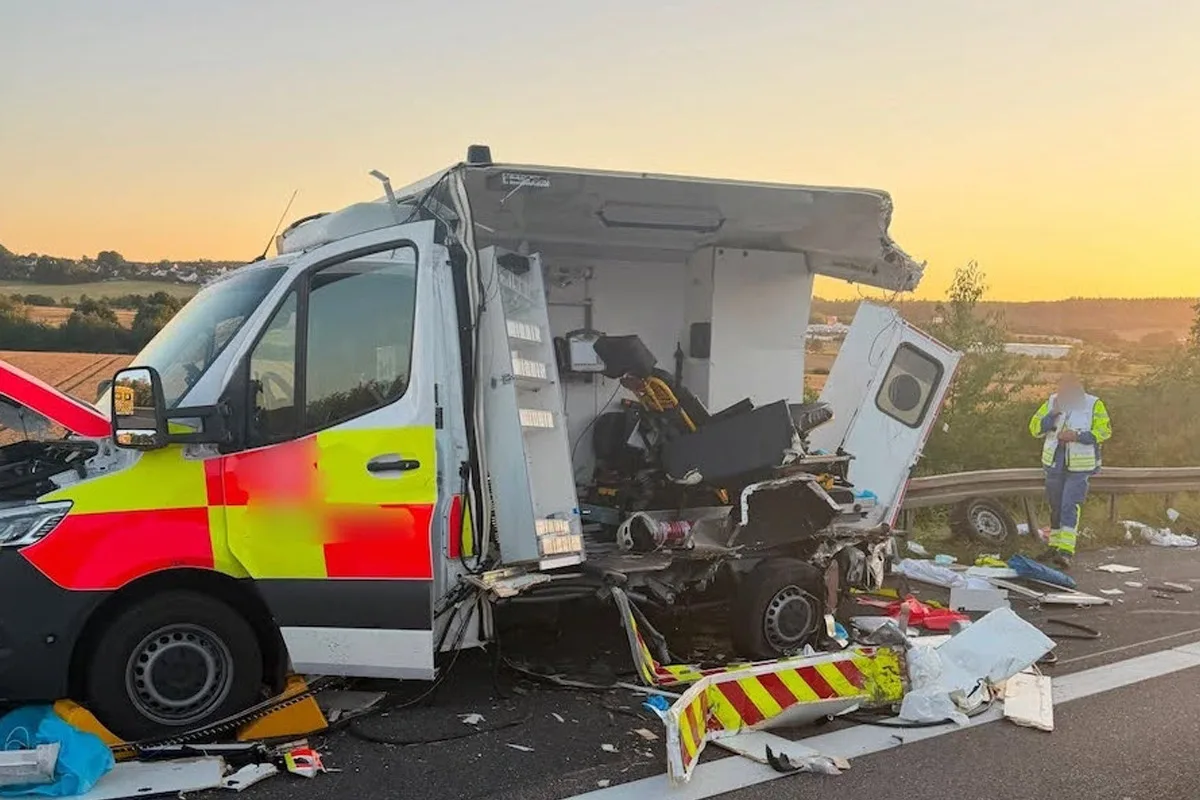 Das Bild zeigt einen schwer beschädigten Rettungswagen auf der Autobahn bei Tagesanbruch. Die Hinterseite des Fahrzeugs ist stark demoliert, während Trümmerteile auf der Straße liegen. Im Hintergrund ist ein Einsatzpersonal in Warnkleidung sichtbar.
