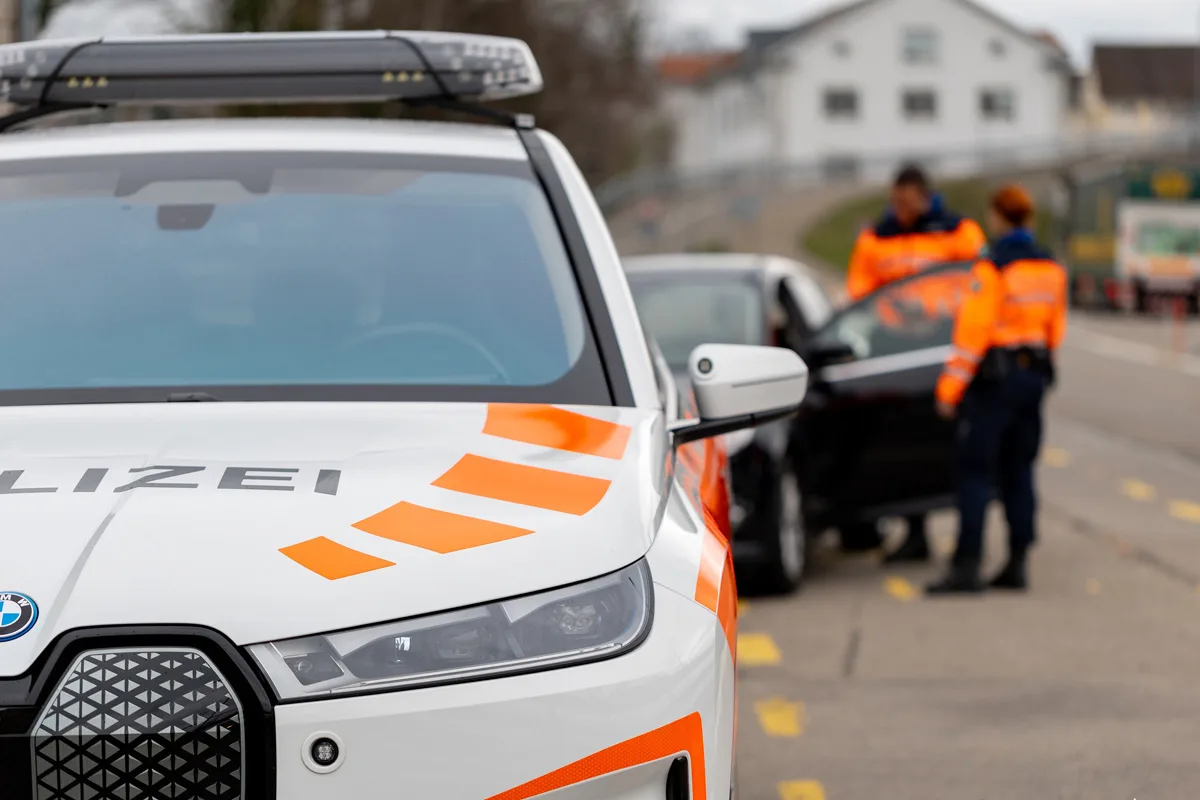 Ein weißes Polizeifahrzeug mit orangefarbenen Markierungen steht im Vordergrund. Im Hintergrund inspizieren zwei Polizisten in orange-blauer Uniform ein anderes Fahrzeug.
