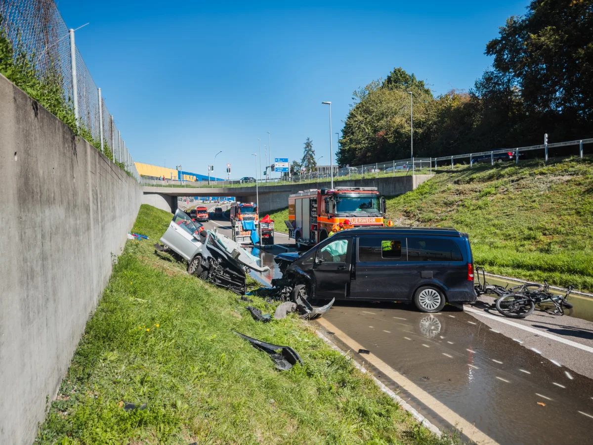 Ein schwarzer Van und ein heller Pkw sind auf einer Autobahnausfahrt kollidiert. Feuerwehrfahrzeuge sind vor Ort.