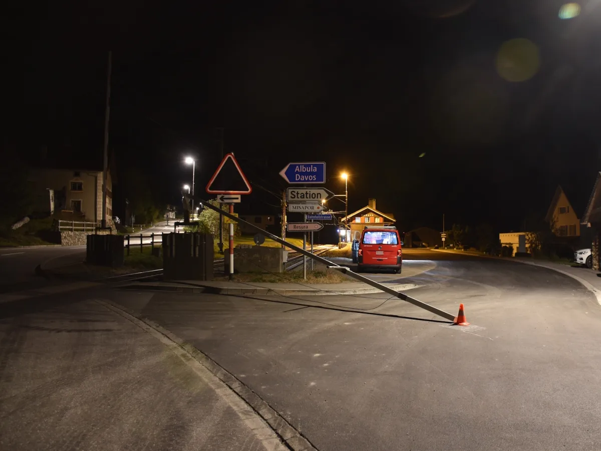 Ein rotes Fahrzeug steht an einer nächtlichen Kreuzung, neben einem umgekippten Verkehrsschild. Die Straßenszene ist schwach beleuchtet, und Häuser sind im Hintergrund sichtbar.